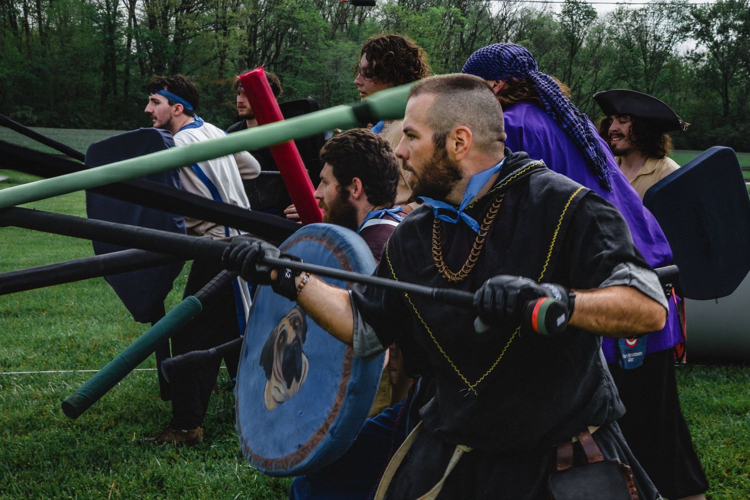 Medieval reenactors in costume engage in a spear combat battle on a grassy field during a live-action roleplay event.