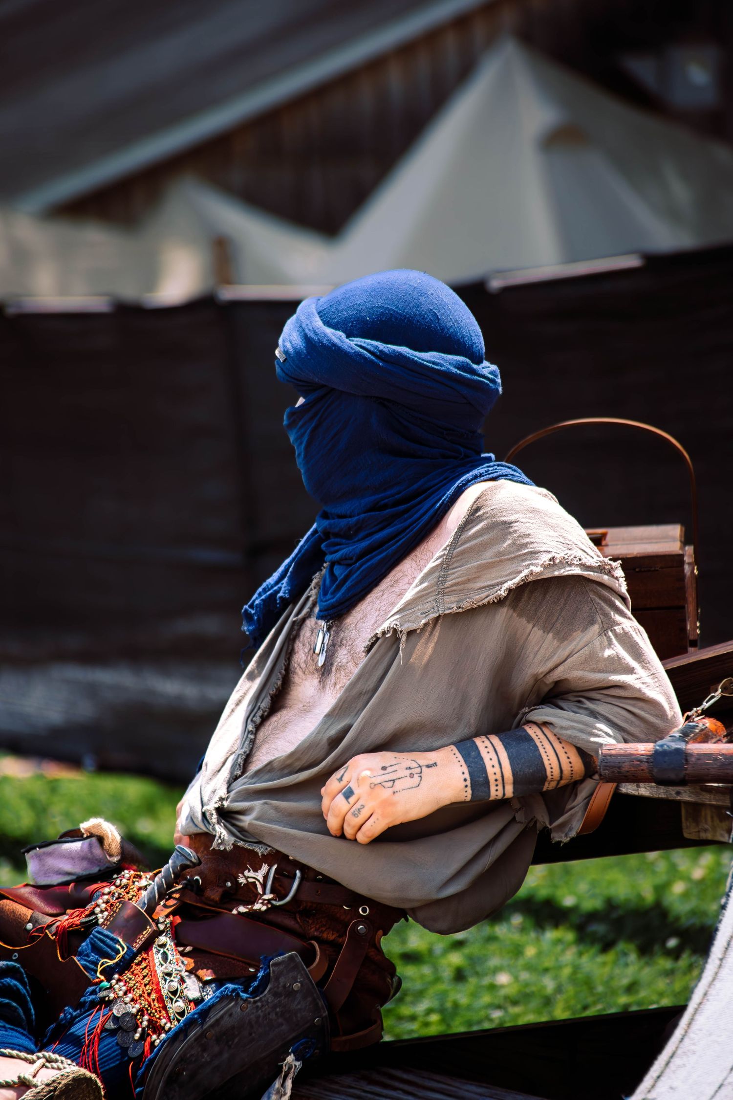 A person in a blue head covering and beige jacket lies sleeping on a wooden bench in a park setting.