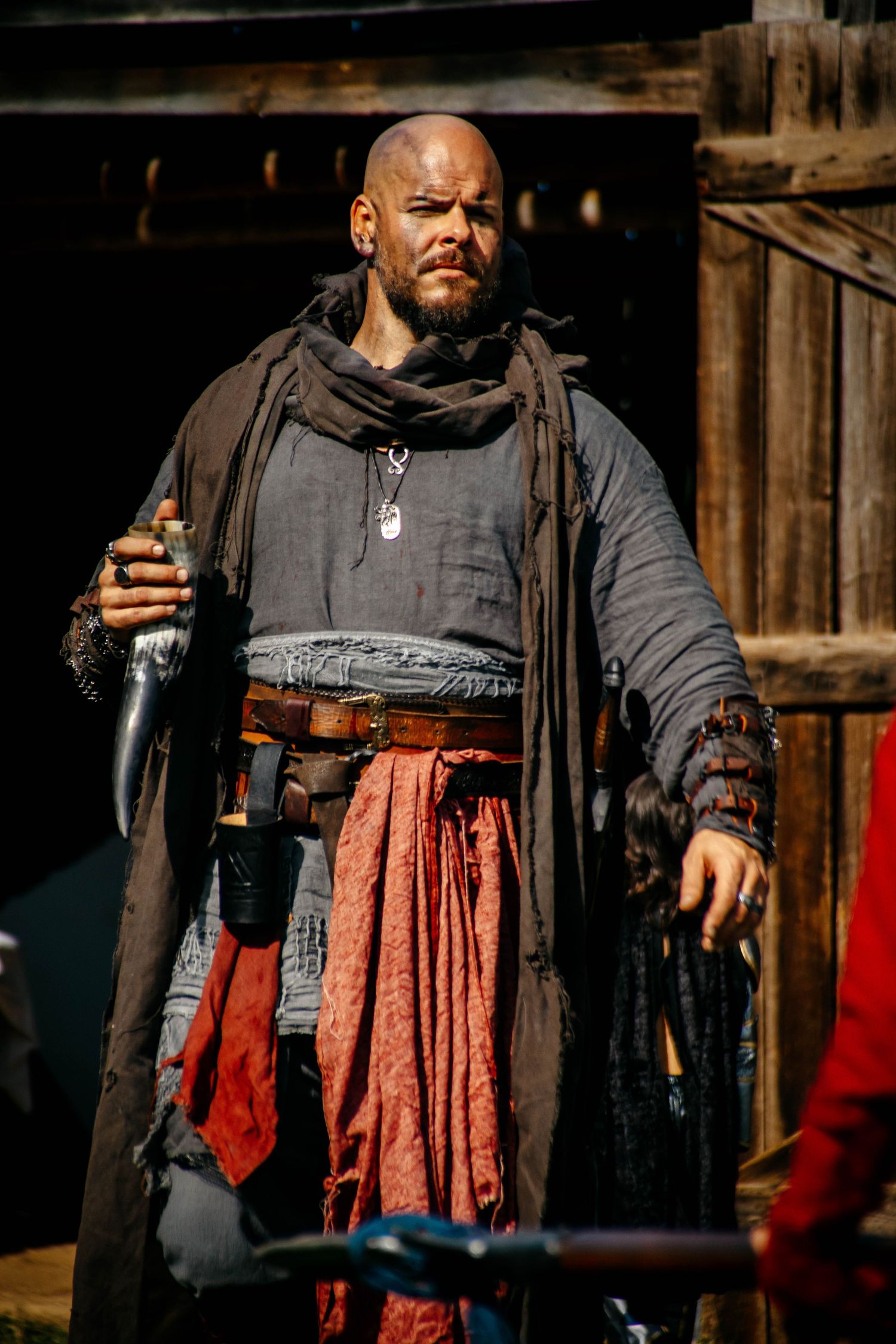 A figure in medieval costume wearing a black vest and red kilt stands with weapons in a rustic wooden setting.
