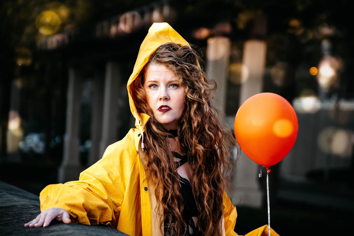 Woman in bright yellow raincoat dances playfully with orange balloons on a cloudy autumn day.
