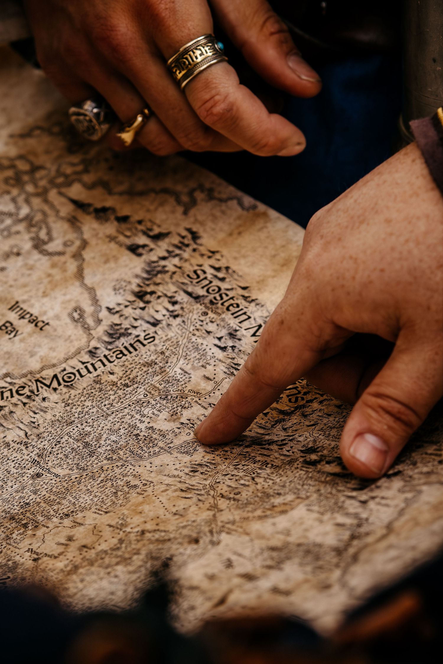 Two hands with rings pointing at an aged antique treasure map with mountain illustrations and old text.