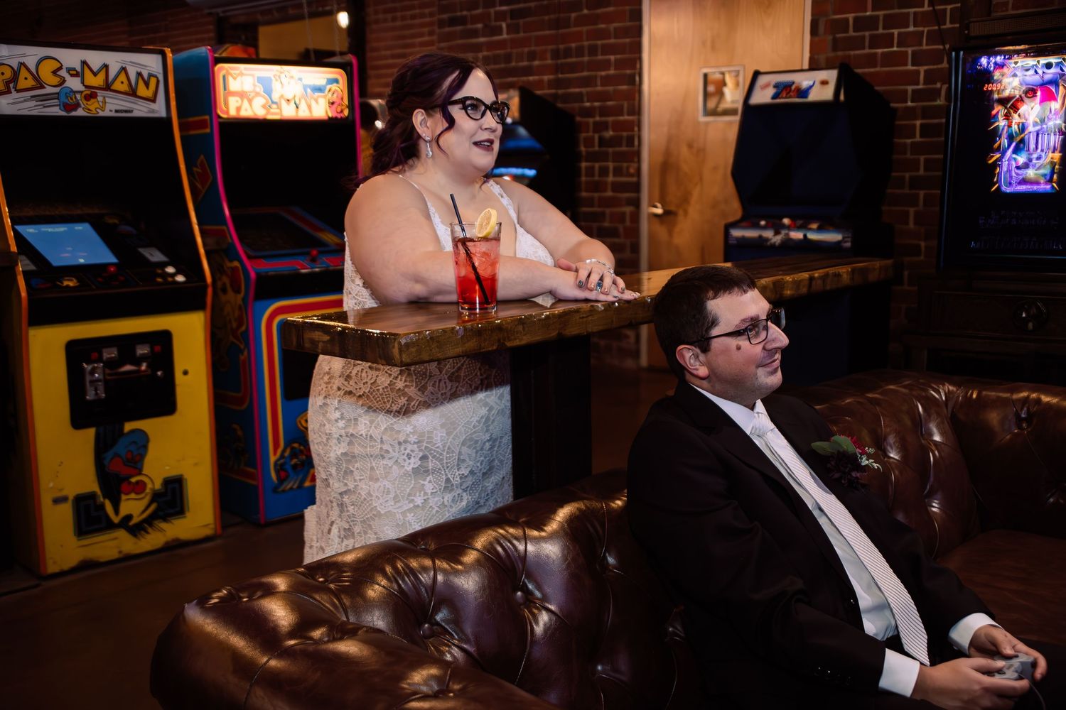 Couple enjoying arcade bar, woman standing at counter near Pac-Man machine, man playing video game on leather couch.
