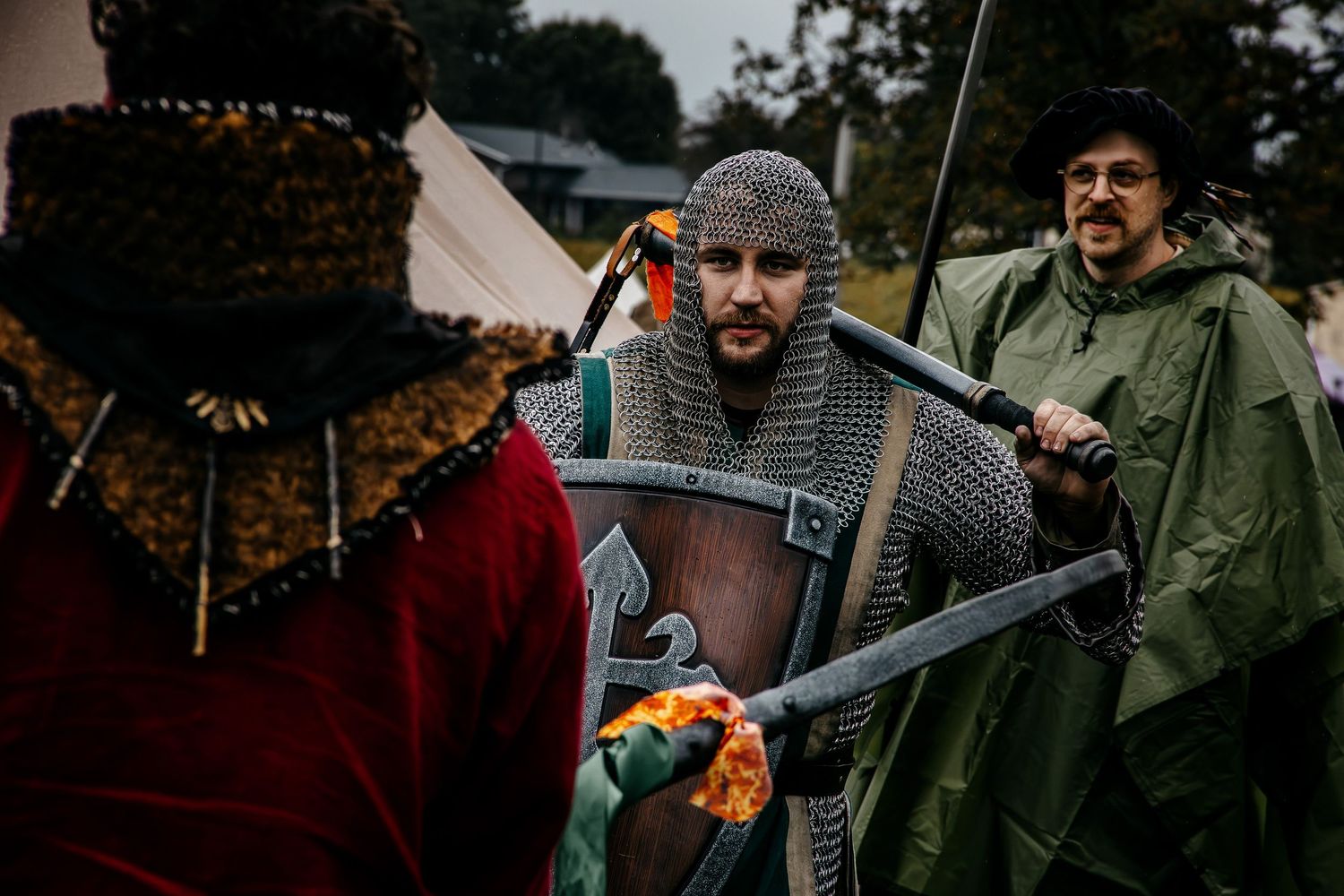 Medieval reenactors in chainmail and period costumes wielding swords and shields at an outdoor historical fair.