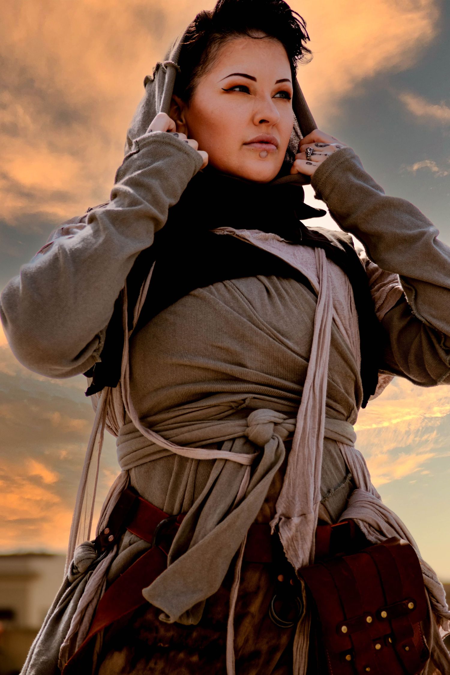A person in medieval-inspired costume with a tan cloak adjusts their headpiece against a dramatic sunset sky.