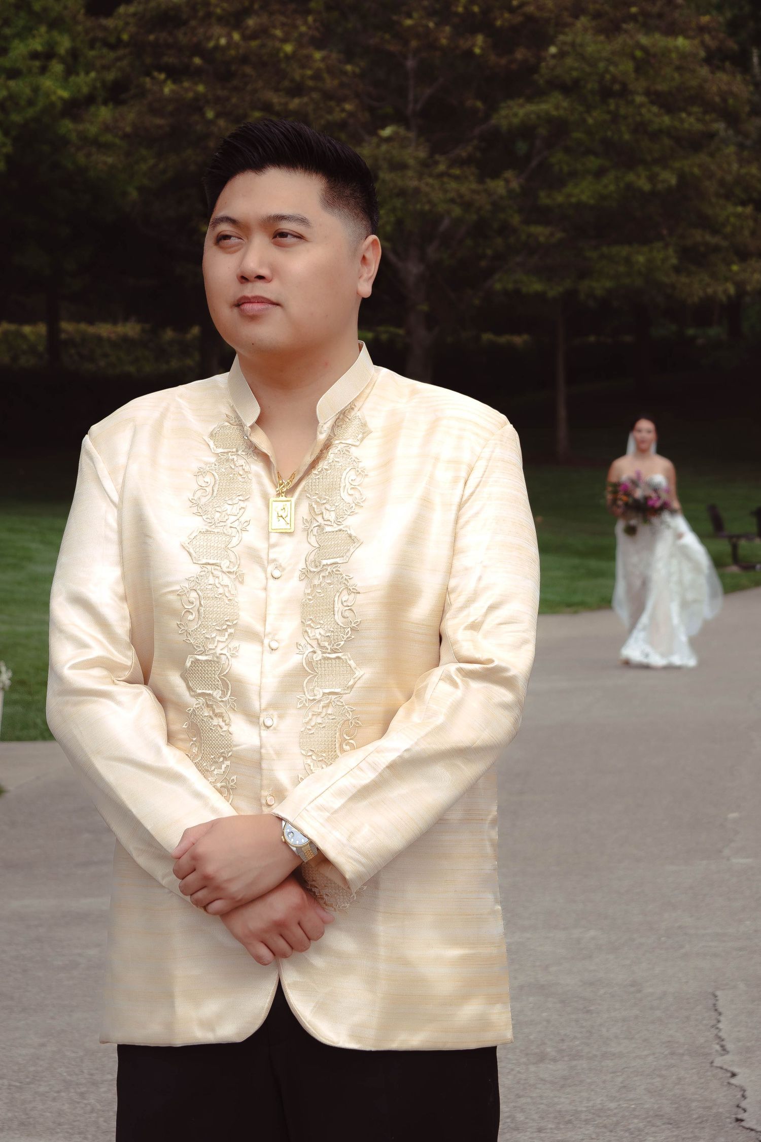 A person in a cream embroidered barong stands on a path while a bride in white is visible in the background.