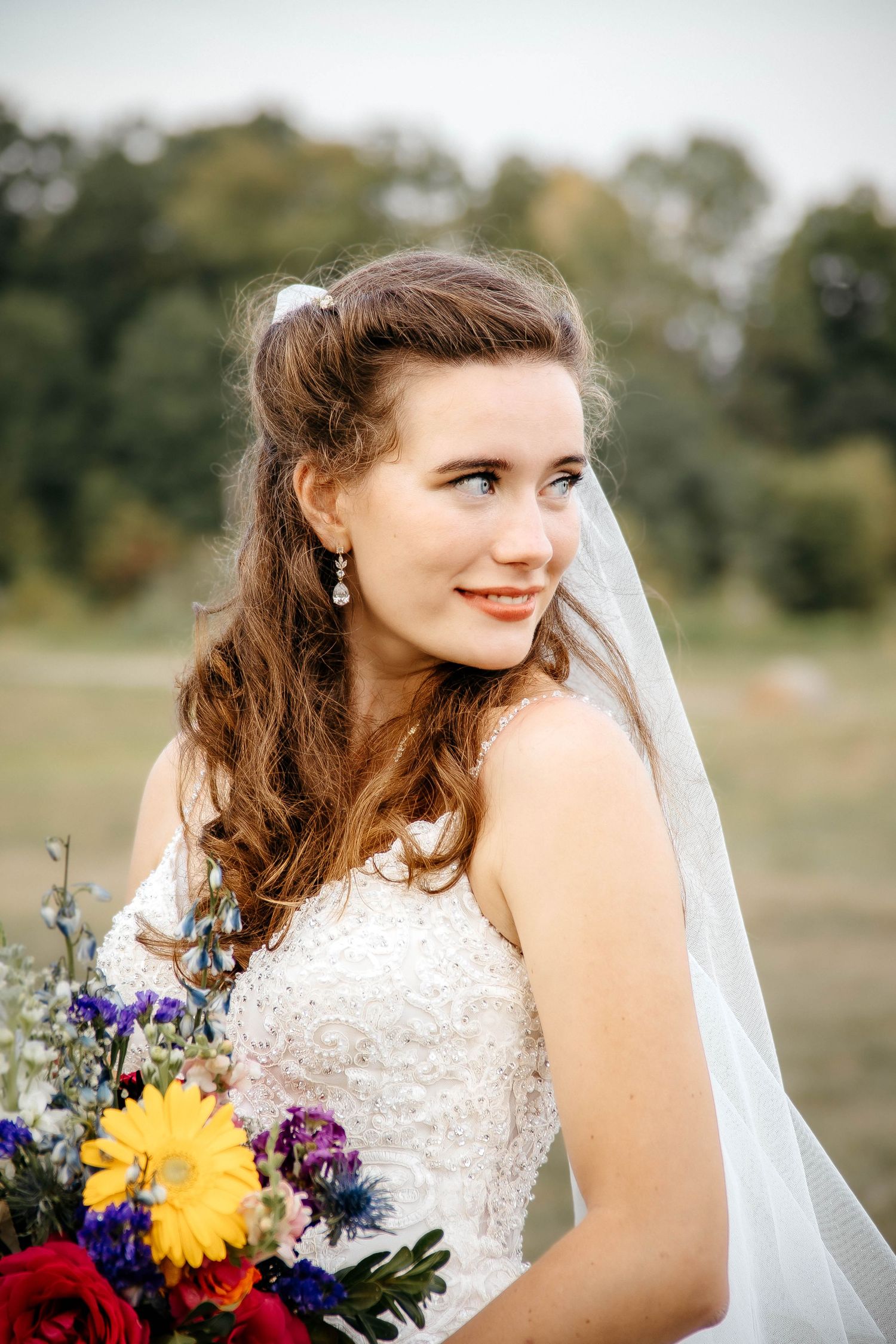 Bride in lace dress holds vibrant sunflower and wildflower bouquet while wearing flowing veil outdoors.