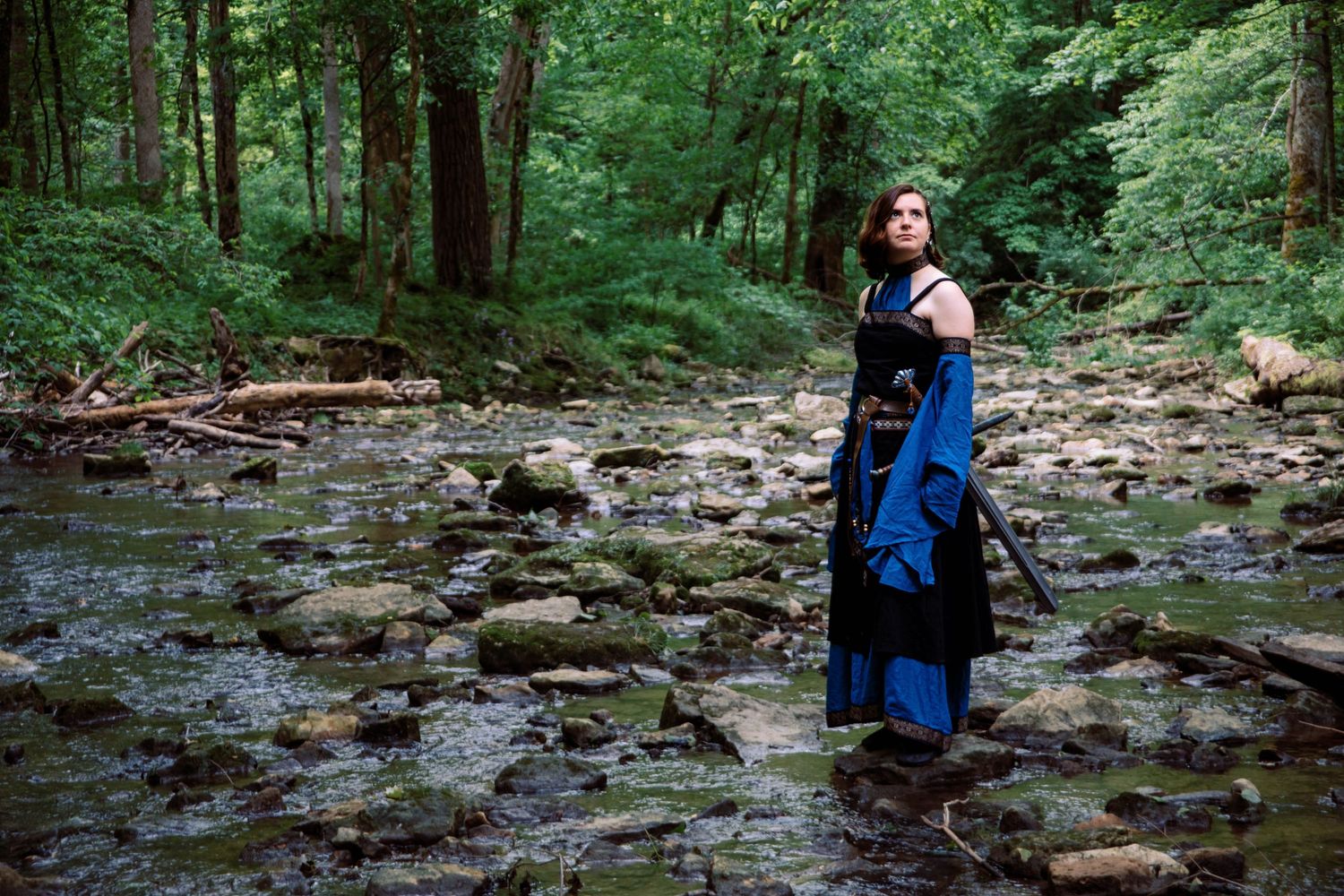 A person in a blue dress stands in a rocky creek bed surrounded by lush green forest.