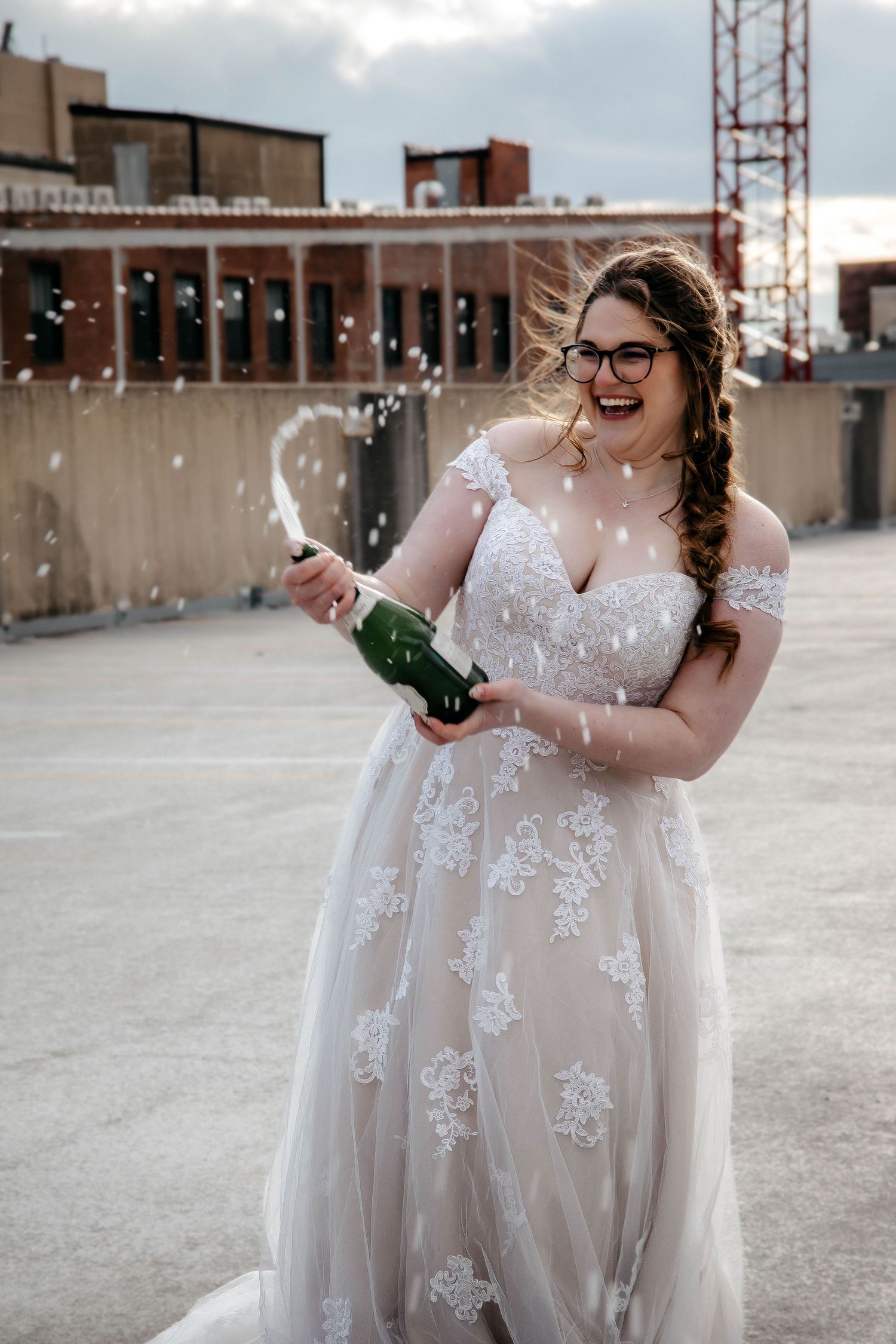 A bride in a lace wedding dress pops open a bottle of champagne on an urban rooftop.