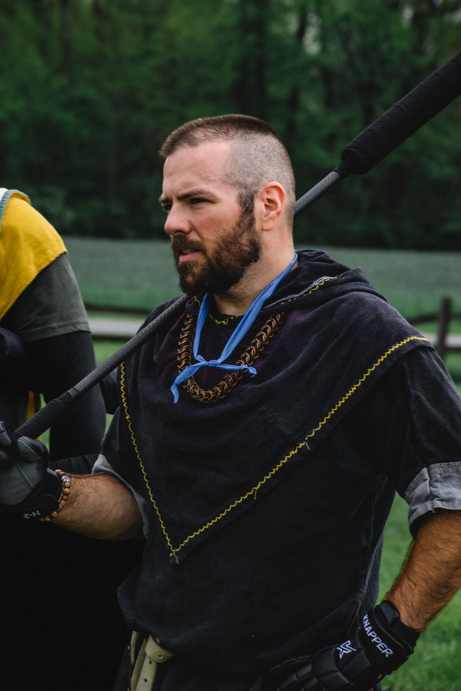 Bearded man in medieval costume holding a polearm weapon at a Renaissance faire event outdoors.