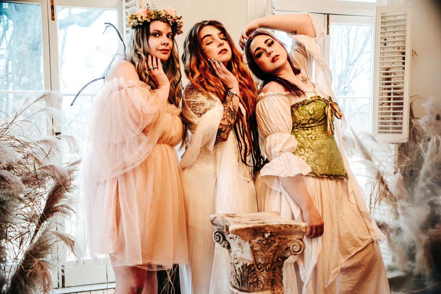 Three women in fairy tale costumes with floral crowns and flowing dresses pose elegantly indoors.
