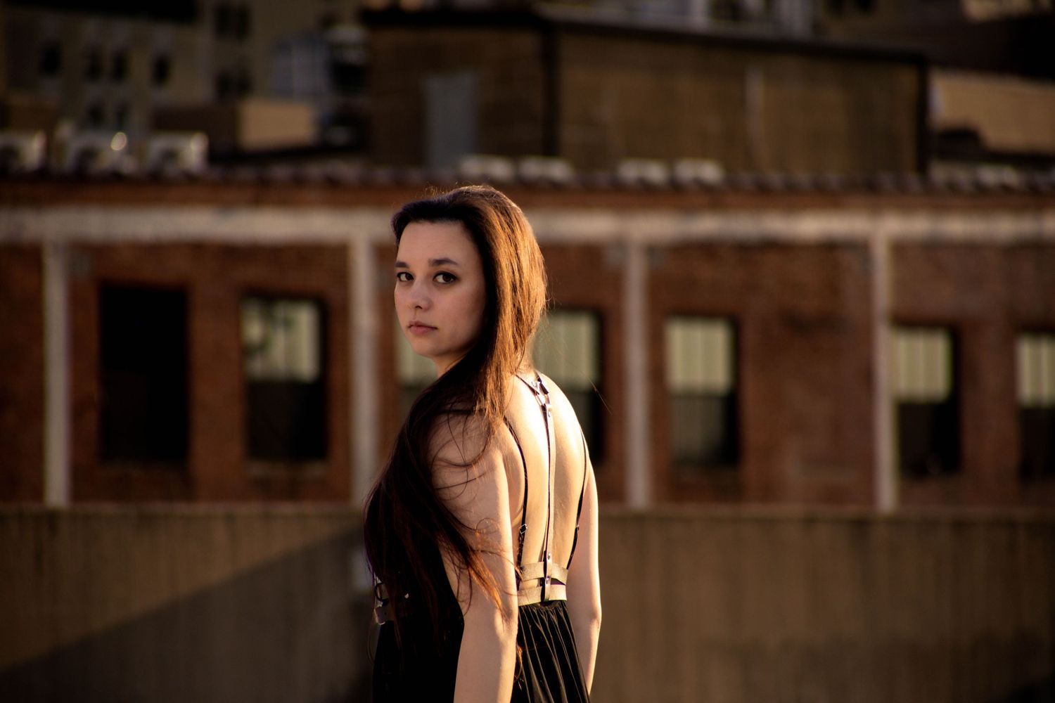 Young woman with long brown hair posing on a rooftop with urban brick buildings in the background.