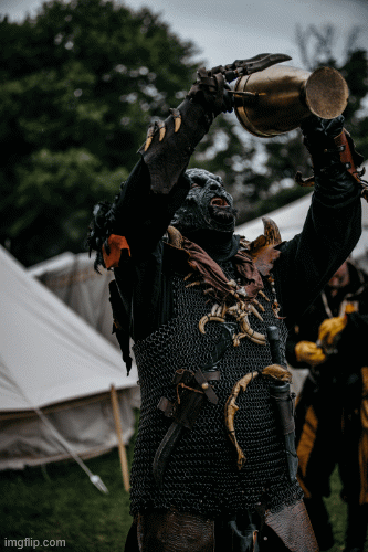 A warrior in chainmail armor and dark fantasy costume raises a horn cup at a medieval LARP reenactment event outdoors.