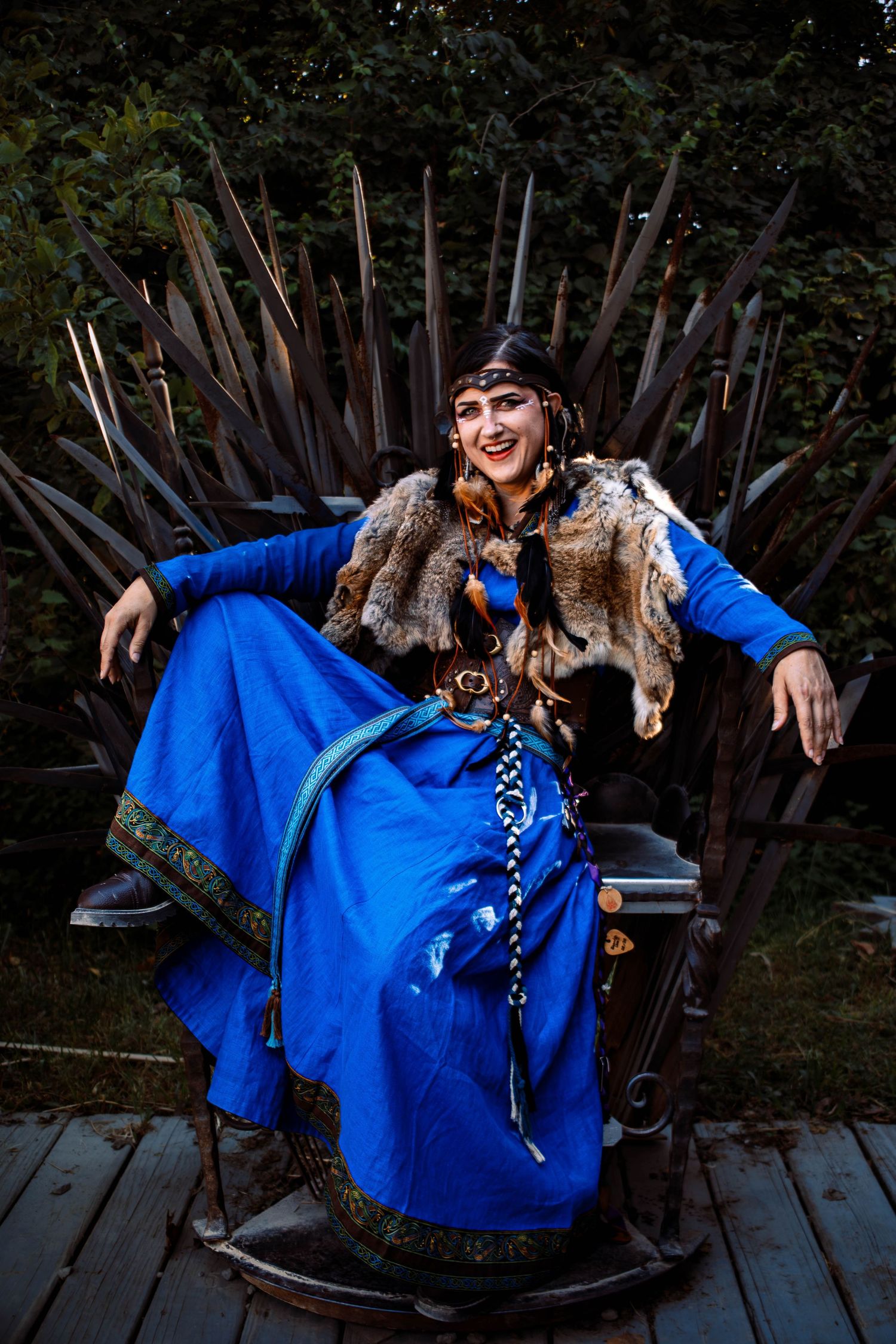 Woman in blue medieval costume with fur vest sits smiling on iron throne replica outdoors.