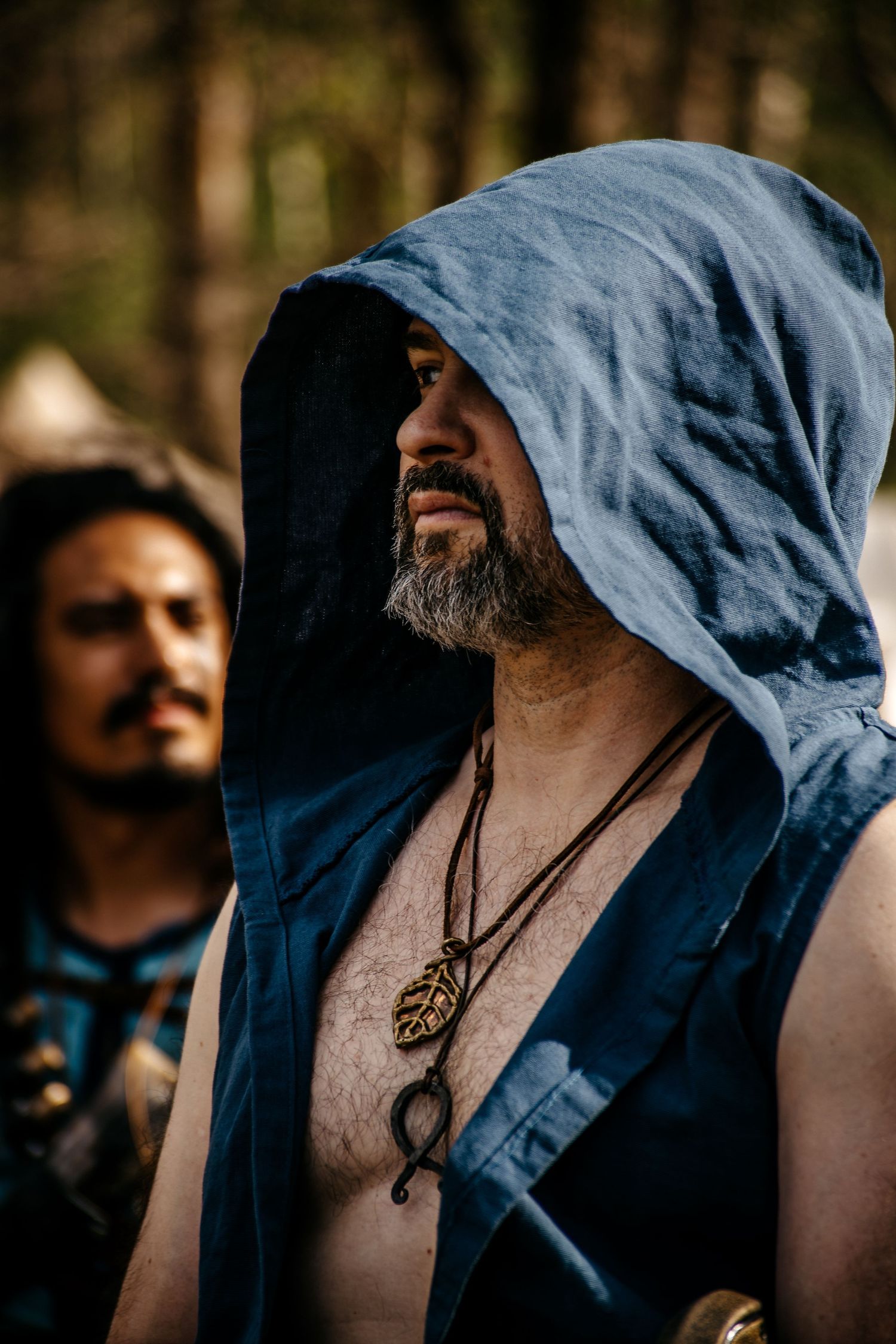 Bearded man in blue hooded medieval vest with leaf pendant necklace at outdoor LARP event.