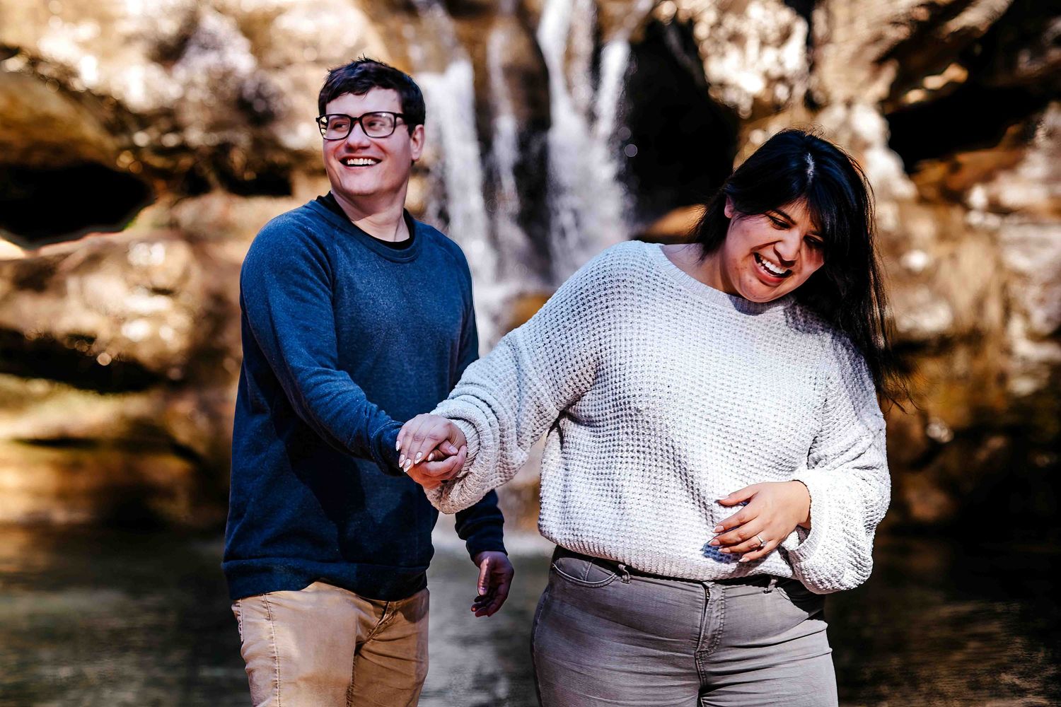 Happy couple holding hands and laughing in front of a waterfall outdoors.