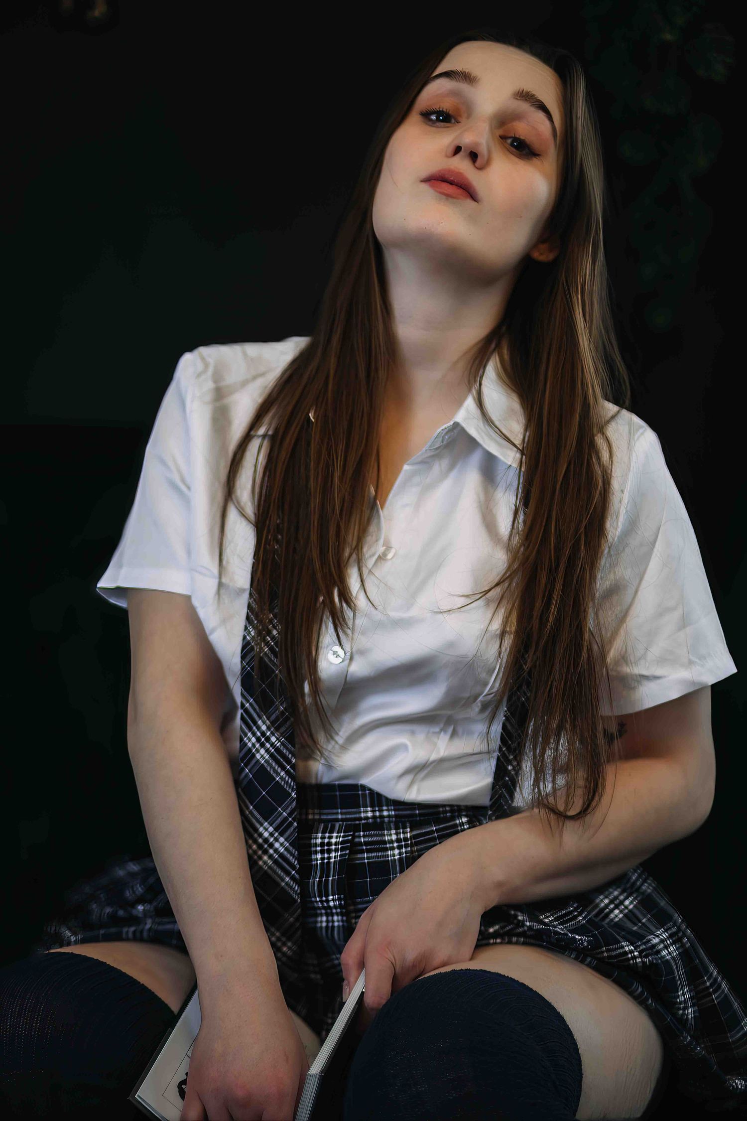 Young woman in white shirt and plaid skirt with bold makeup poses dramatically against dark background.