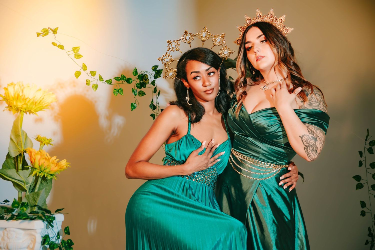 Two women in elegant teal gowns and gold crowns pose gracefully among yellow flowers and green vines.