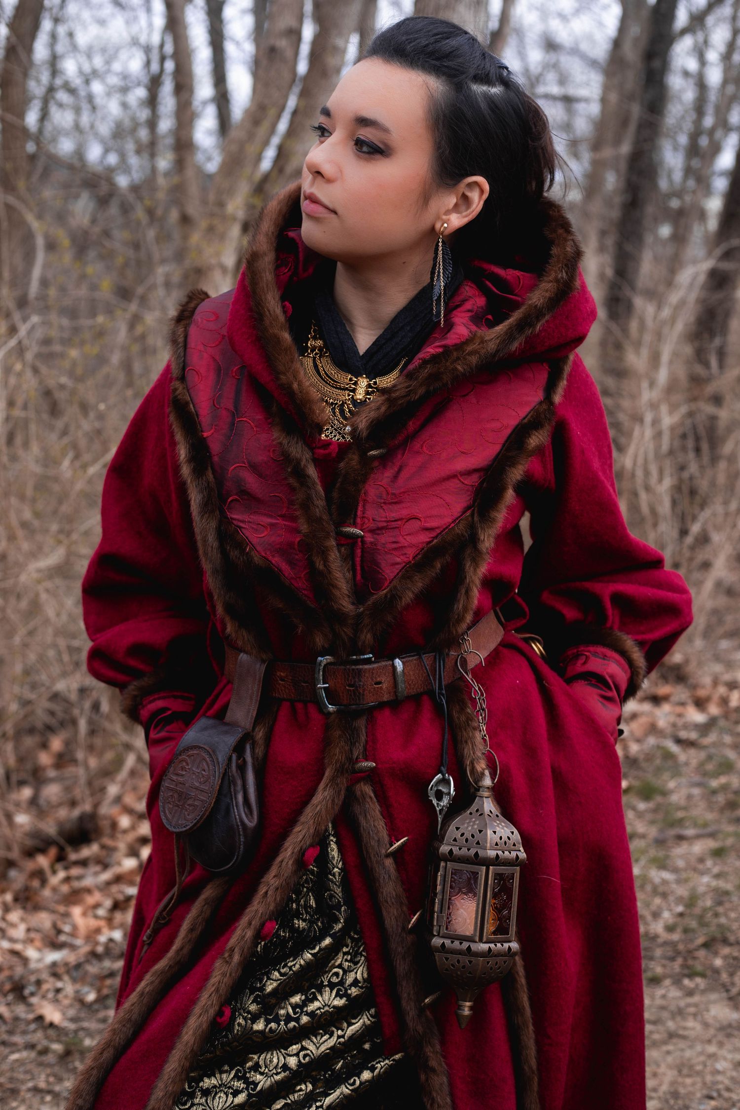 Fantasy medieval costume with deep red hooded cloak, leather belt and decorative lantern in autumn forest setting.