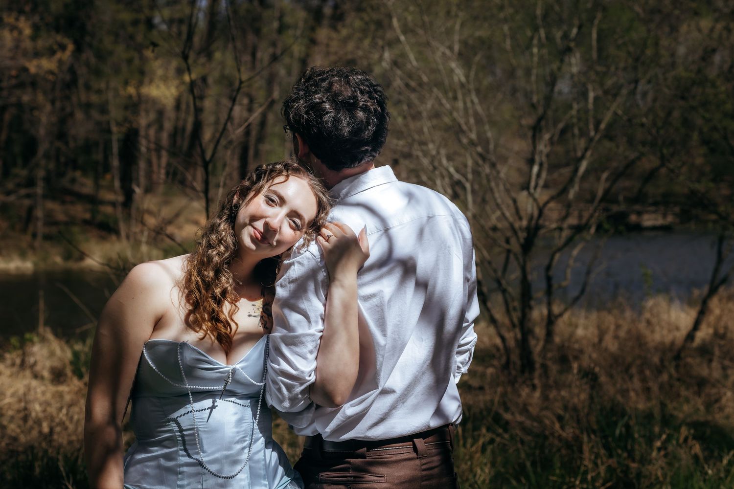 A couple shares a romantic moment outdoors, with the woman in a blue dress smiling warmly at her partner near a forest lake.