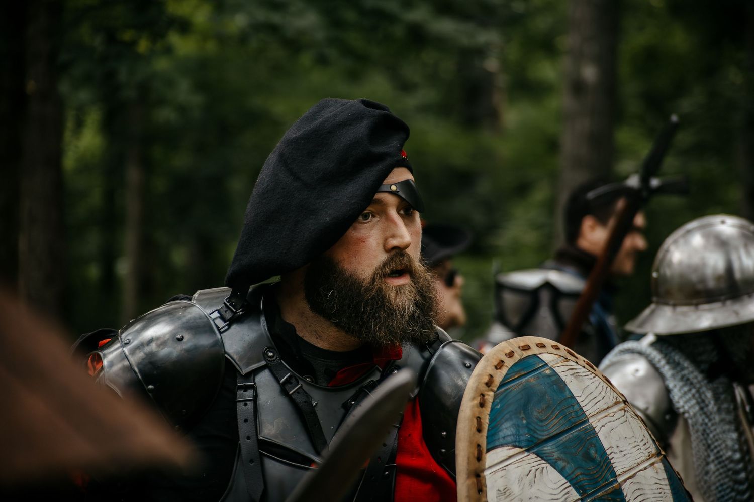 Bearded medieval knight in black hood and armor holding shield during forest battle reenactment.