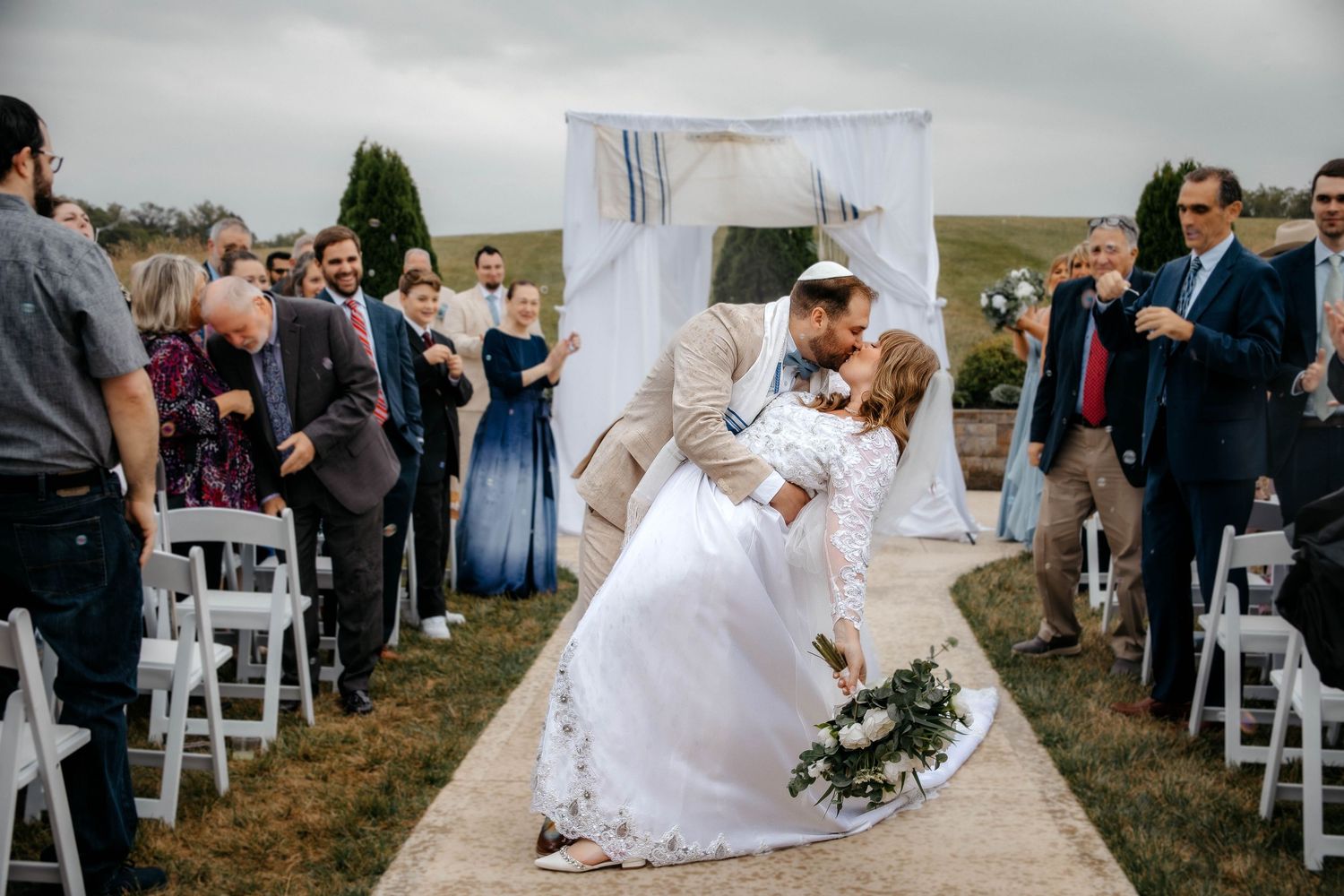 A wedding couple shares an intimate kiss during their outdoor ceremony while guests look on from white chairs.