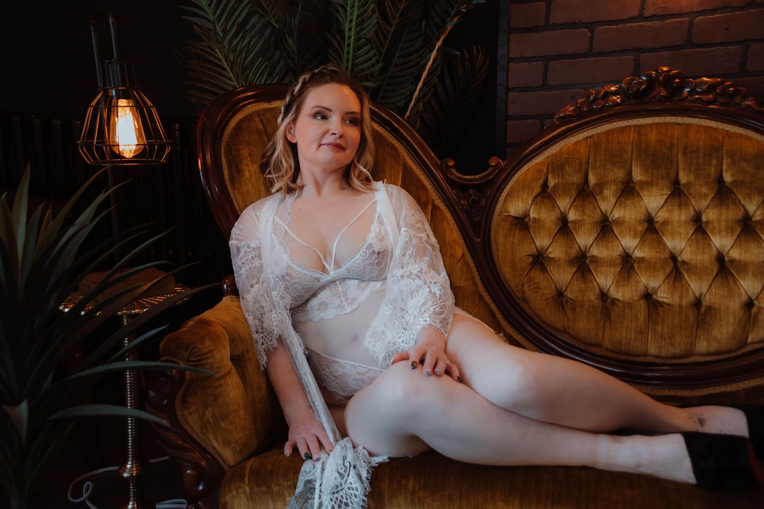 Elegant figure in white lace robe reclines on vintage gold velvet sofa beneath hanging pendant light and palm fronds.