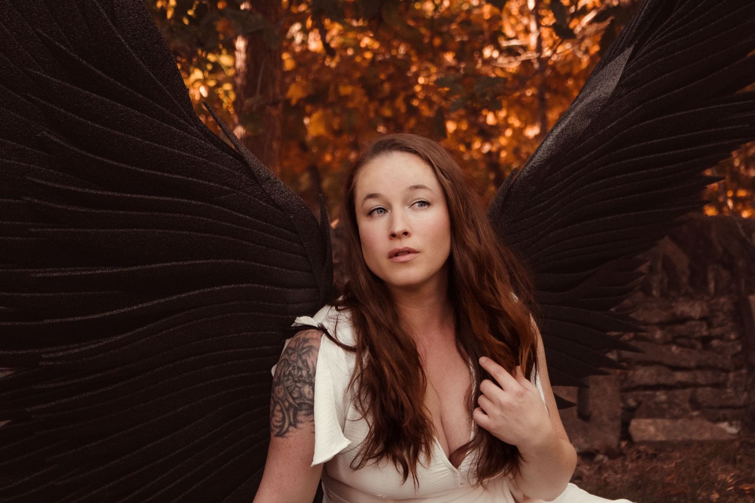 A figure in a white dress with dark wings poses against a warm autumn background with brick wall details.