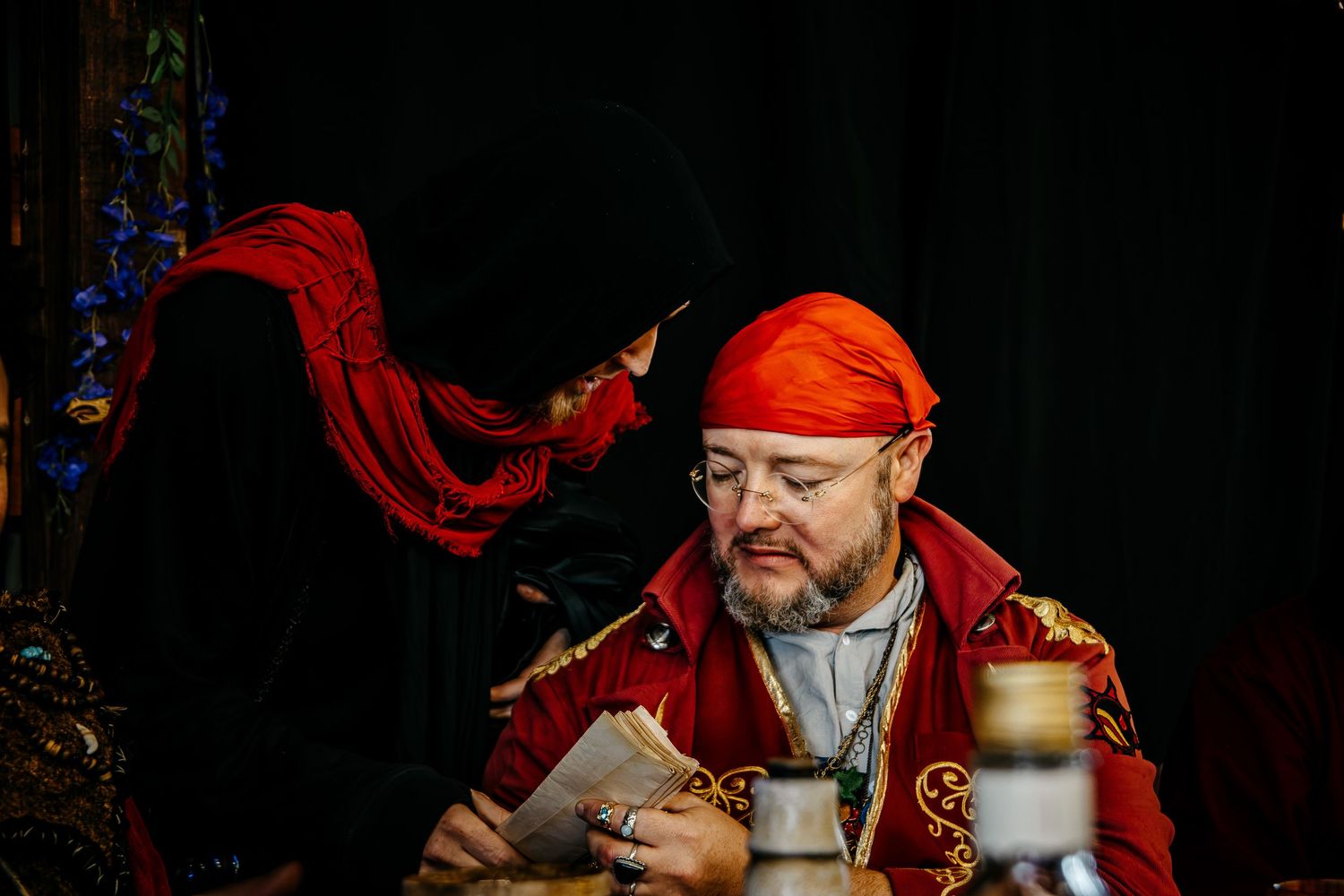 Two actors in elaborate red and black costumes perform a dramatic scene, with one reading a letter in a theatrical setting.