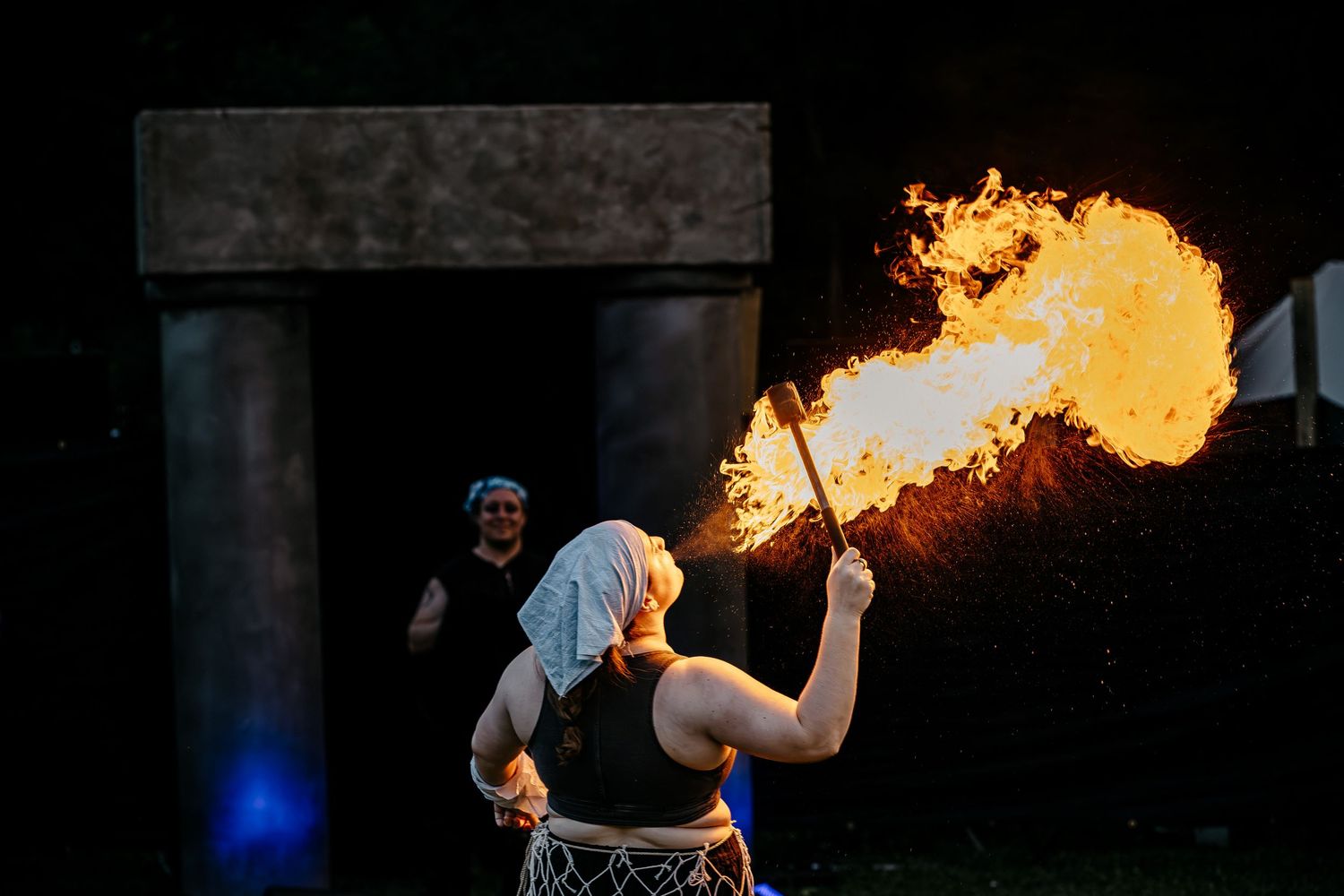 A fire breather performs a dramatic flame stunt at night, with a spectator watching near a stone doorway.