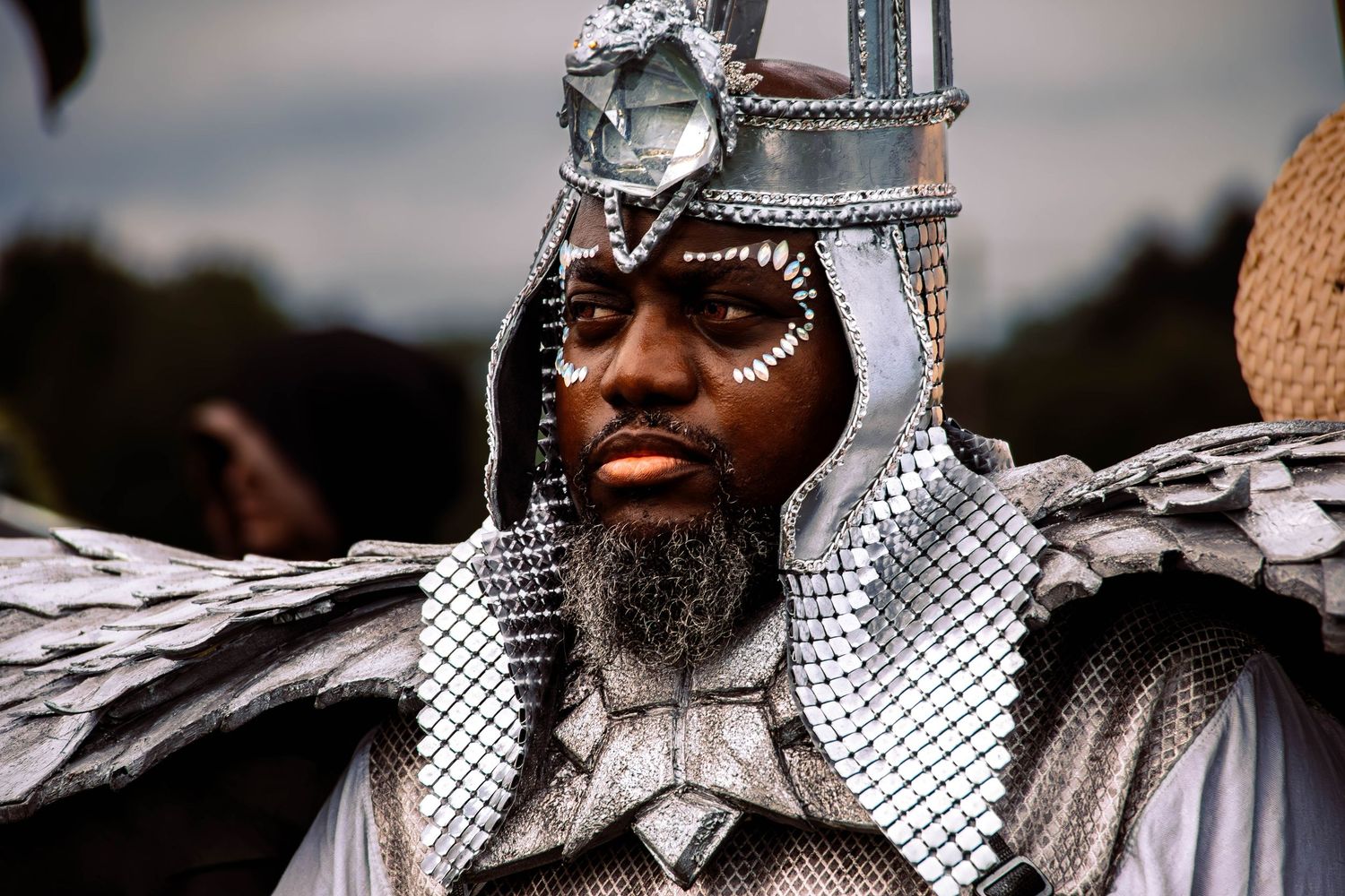 A medieval knight in shining chainmail armor and metallic crown stands regally during a historical reenactment event.