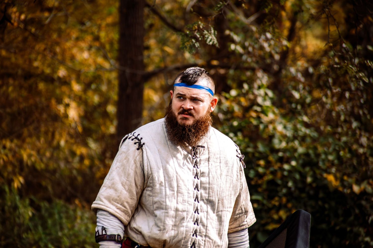 A person wearing a white medieval-style tunic and headband stands in a sunlit autumn forest setting.