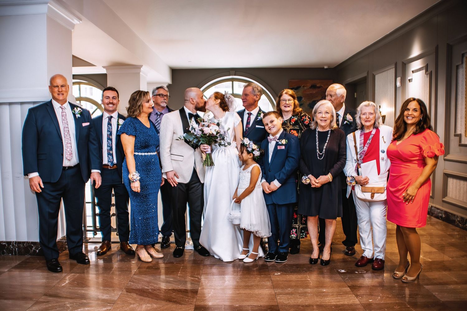 Wedding party poses together indoors, bride and groom kissing at center, surrounded by smiling family members in formal attire.