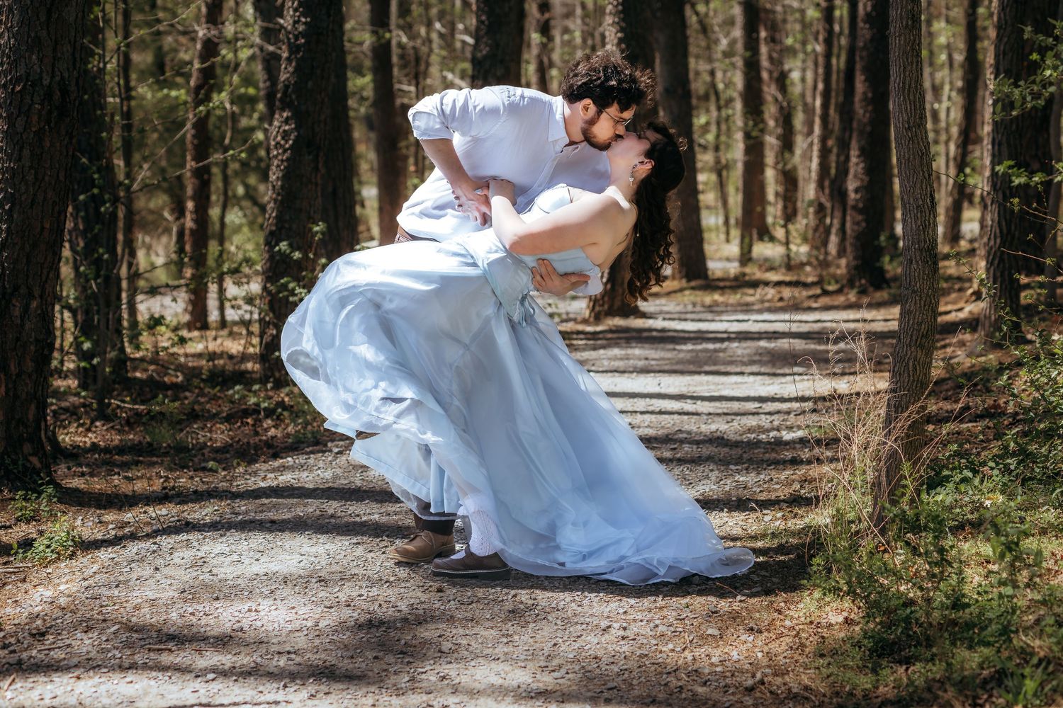 A groom dips his bride for a romantic kiss on a forest path, her white wedding gown flowing gracefully.