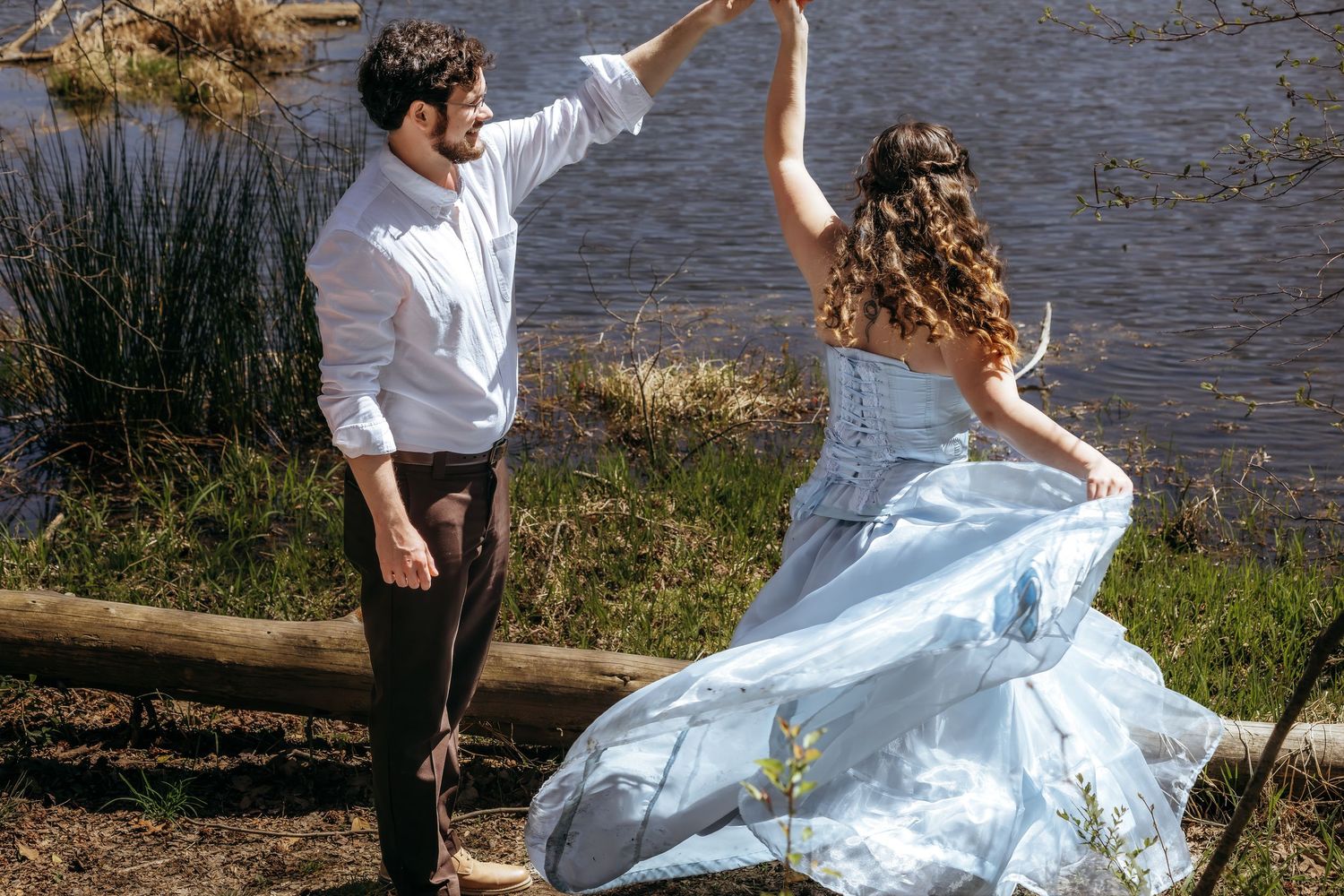 Couple dancing in wedding attire by a lake, bride in flowing light blue gown, groom in white shirt and black pants.