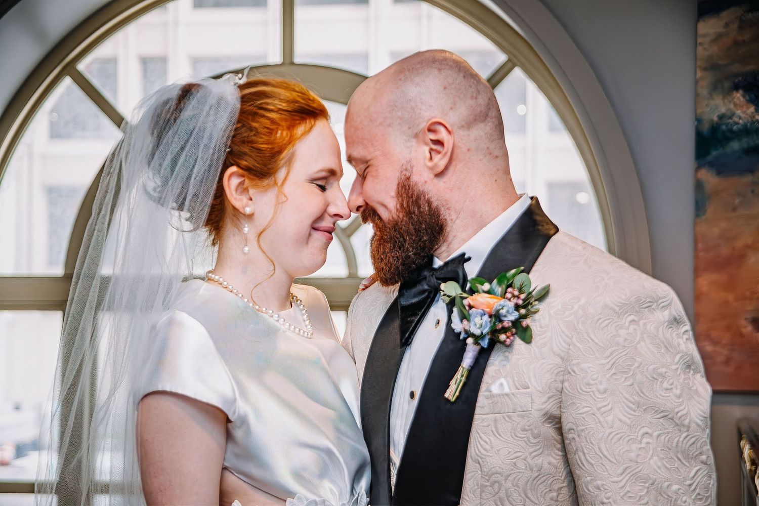 Bride and groom share a romantic moment, touching foreheads, dressed in wedding attire with a floral boutonniere.