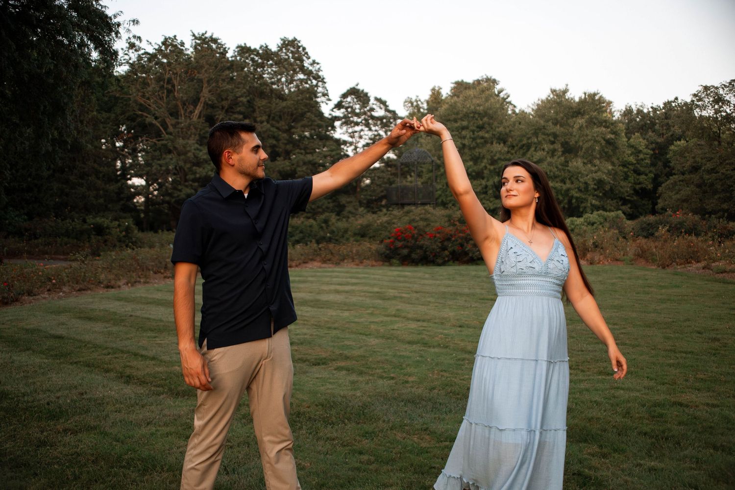 A couple in casual attire dances playfully together in a grassy field during golden hour.
