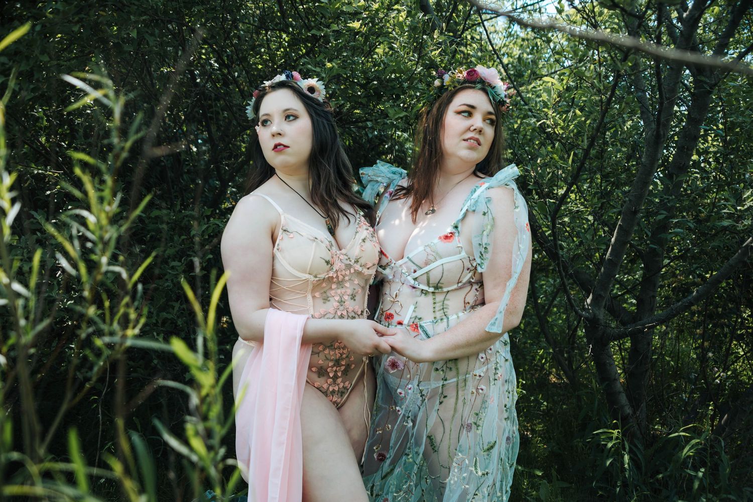 Two people in ethereal dresses pose together in a lush garden setting.