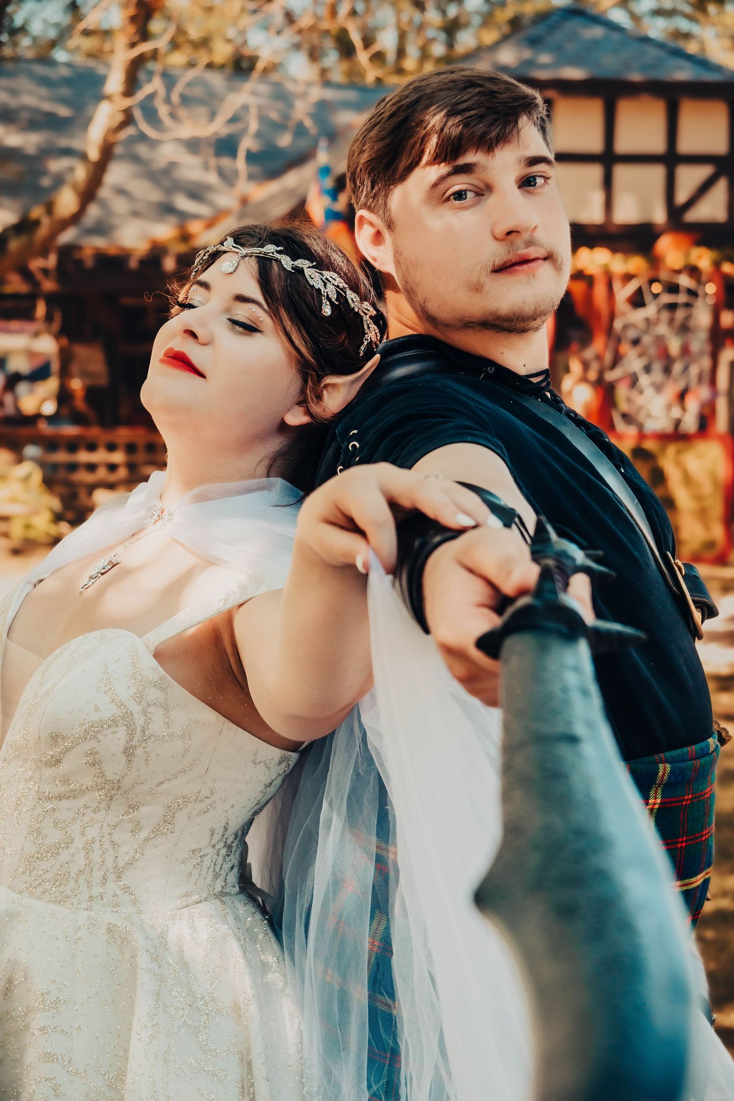 A bride in white dress and groom in Scottish kilt pose back-to-back holding a sword at a Renaissance faire wedding.