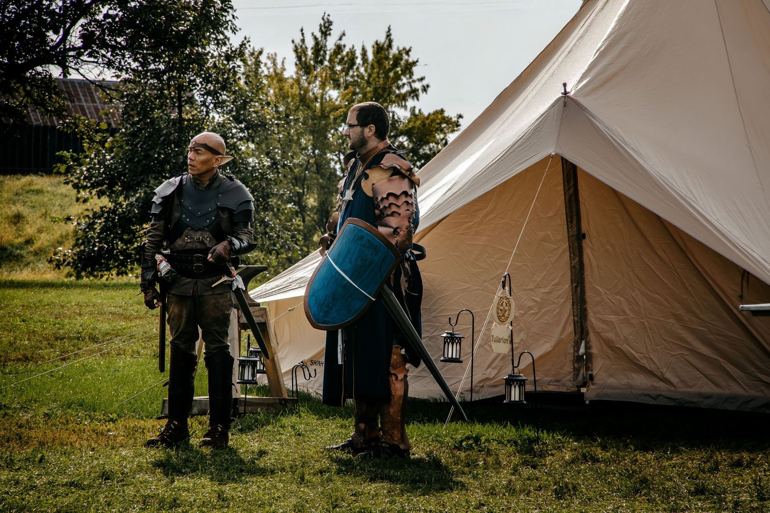 Two medieval reenactors in costume stand near a canvas bell tent on a grassy field at an outdoor event.