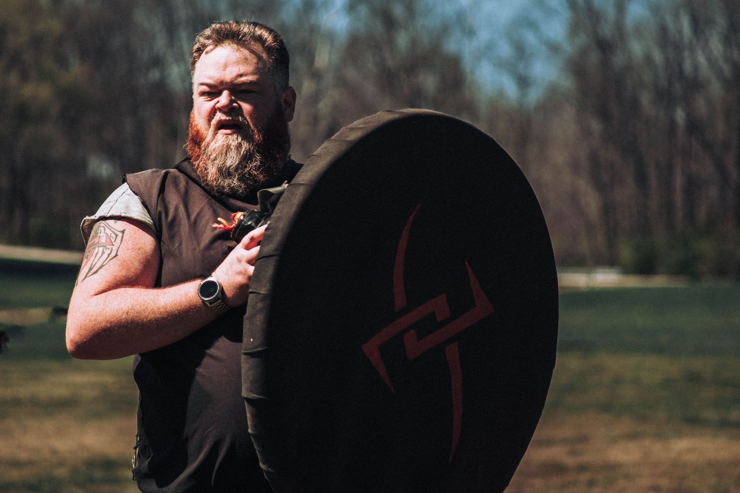 A person practices shield blocking techniques in a grassy outdoor training area on a sunny day.