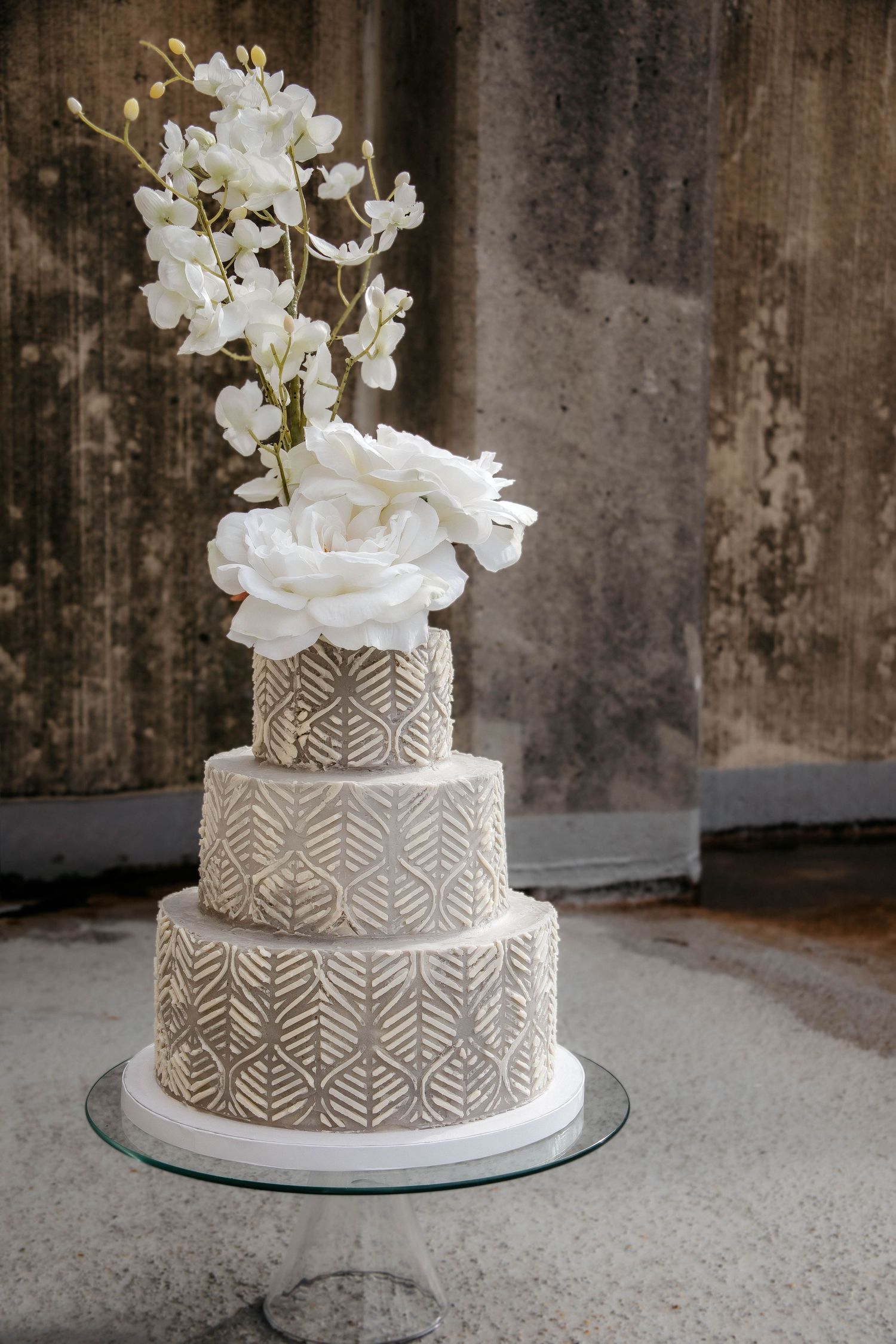 Elegant white wedding cake with geometric pattern and floral topper displayed on glass stand against textured wall.