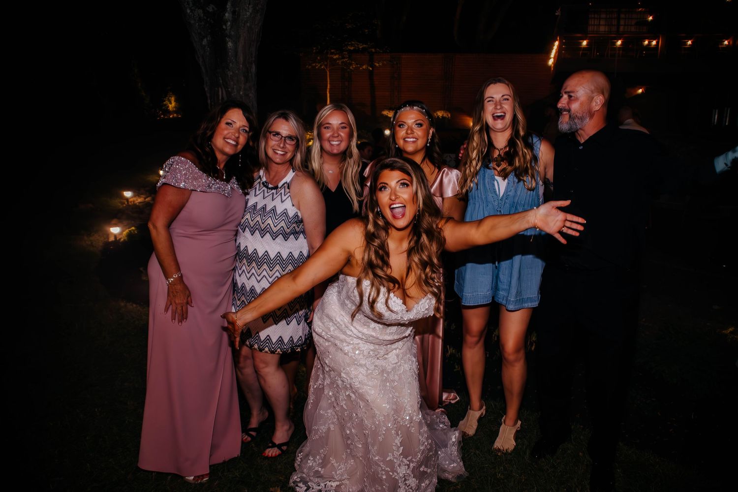 Bride in lace gown laughing with wedding guests outdoors at night during reception celebration.