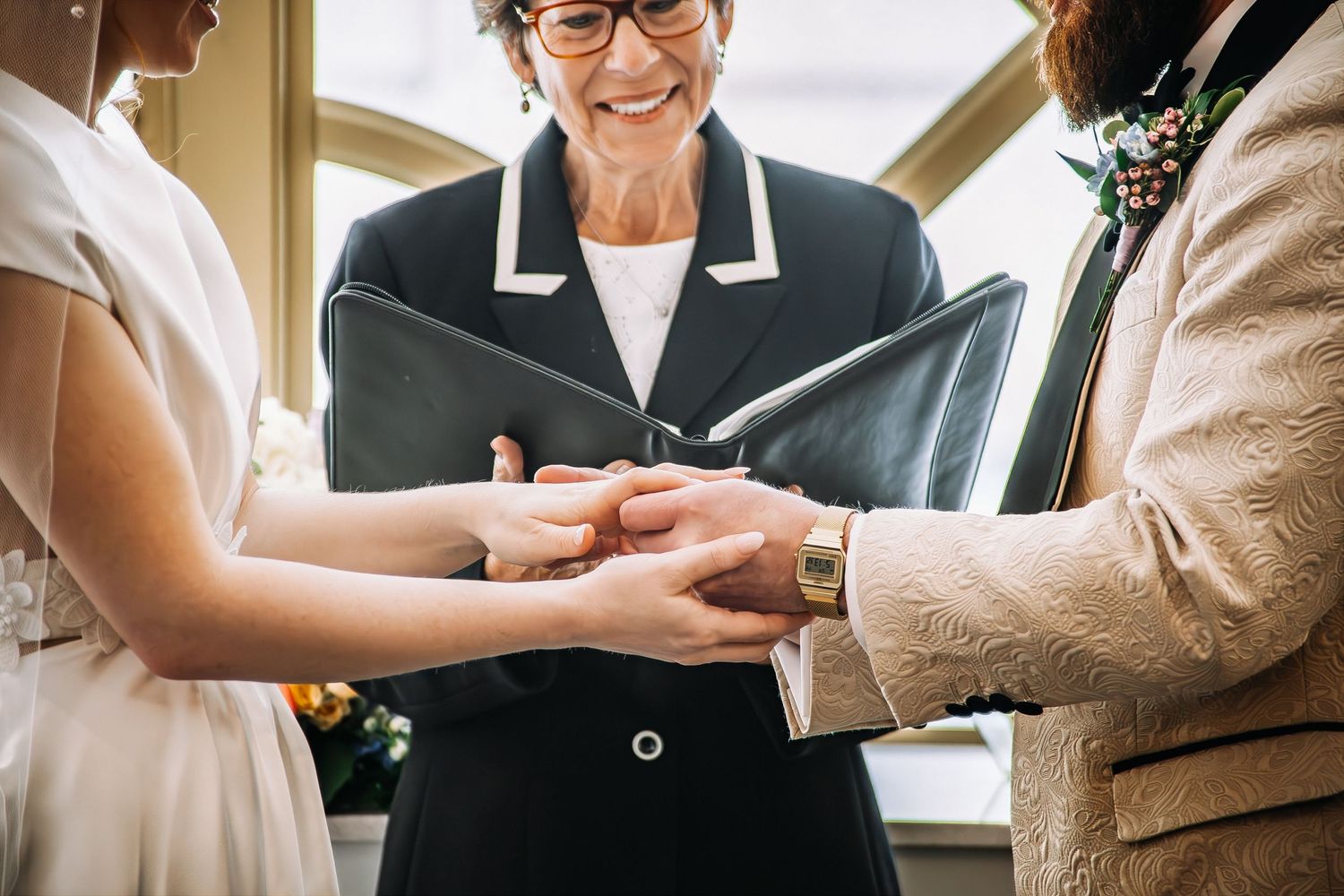 Wedding officiant holds hands of bride and groom during ring exchange ceremony indoors.