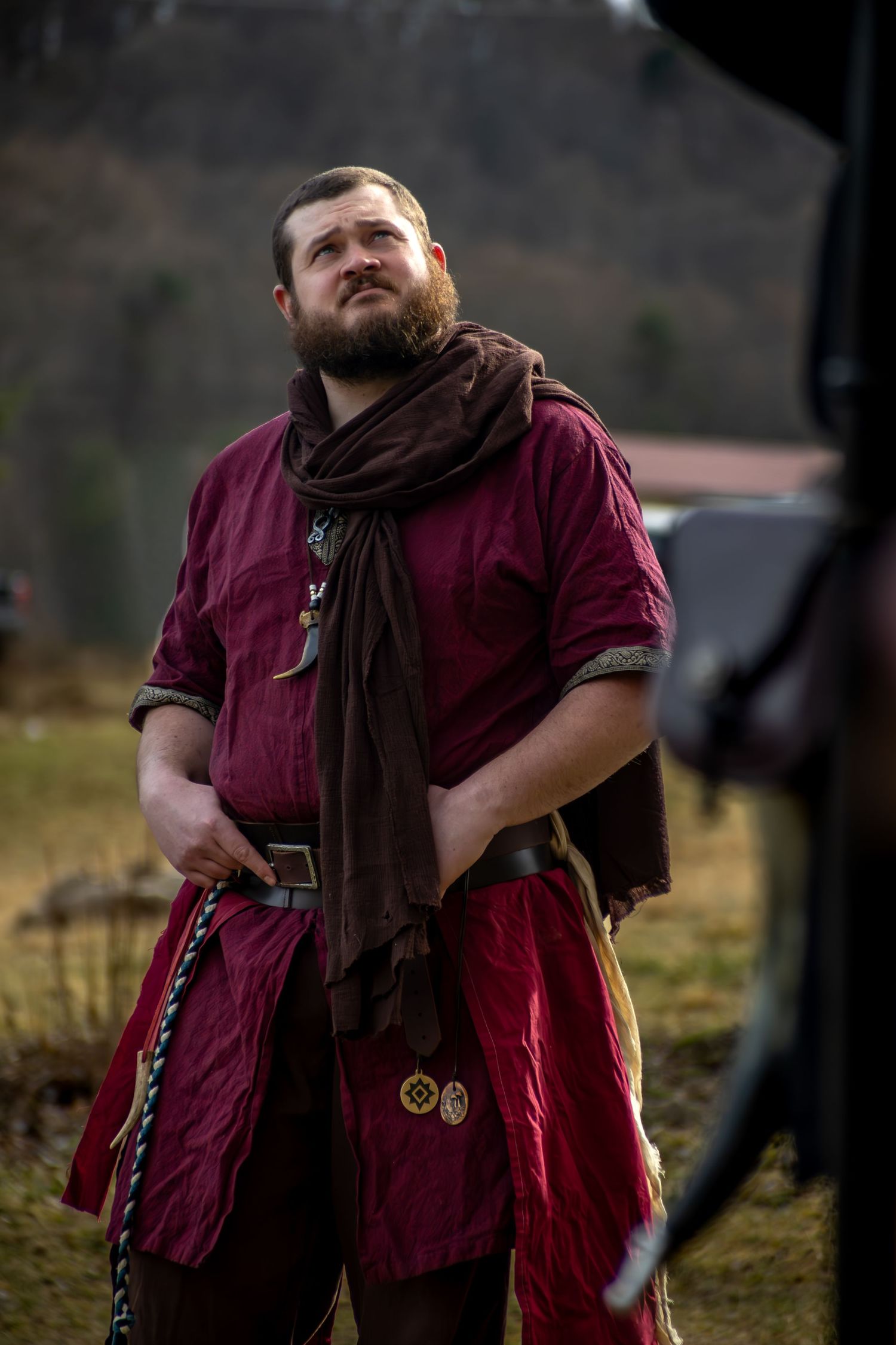 A bearded man in medieval red tunic with brown scarf and belt looks upward outdoors.