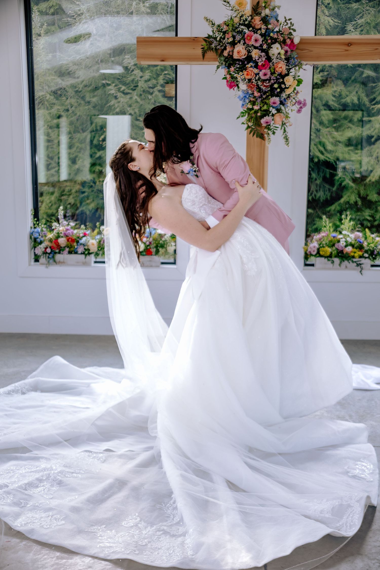Bride and groom share first kiss in elegant wedding venue with floral arch and white bridal gown with long veil.