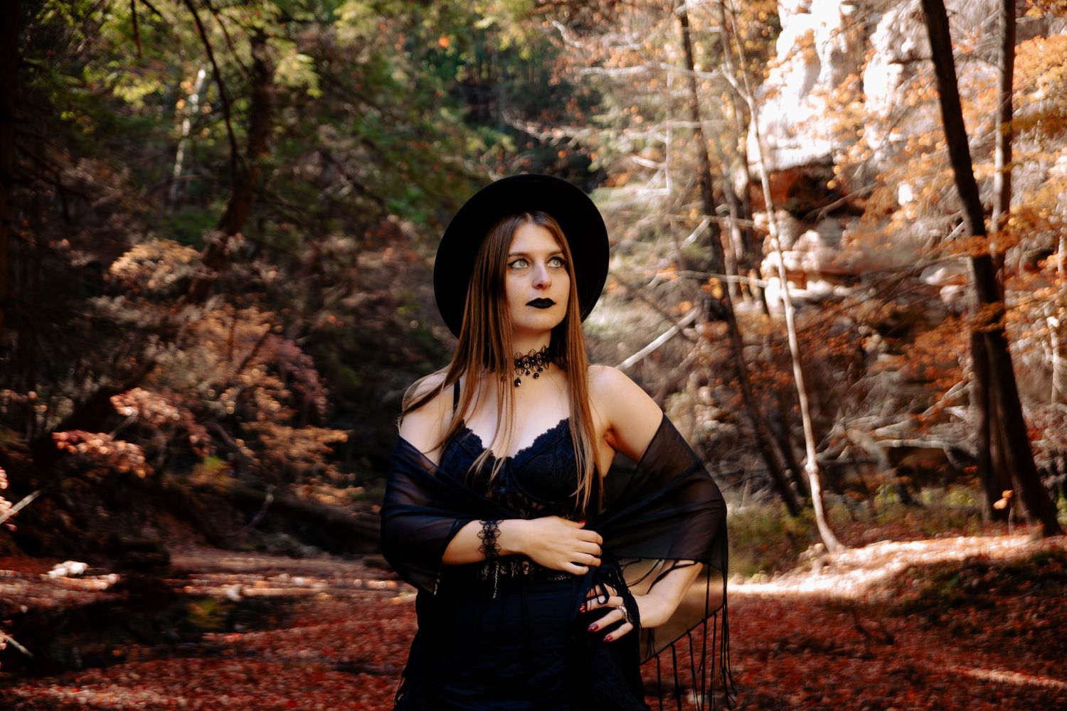 Woman in witch costume with black hat and dark outfit poses in autumn forest with red and orange foliage.