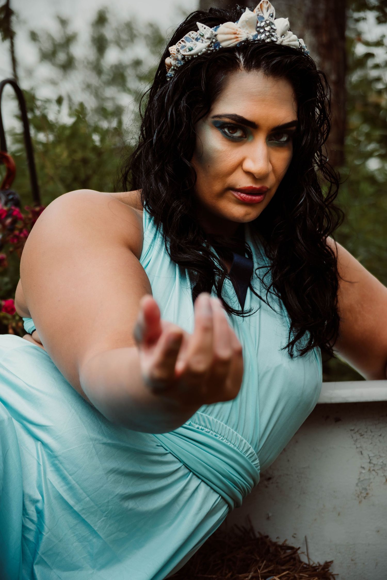 Woman in teal gown and floral crown reaches toward camera with dramatic makeup in an outdoor fantasy portrait.