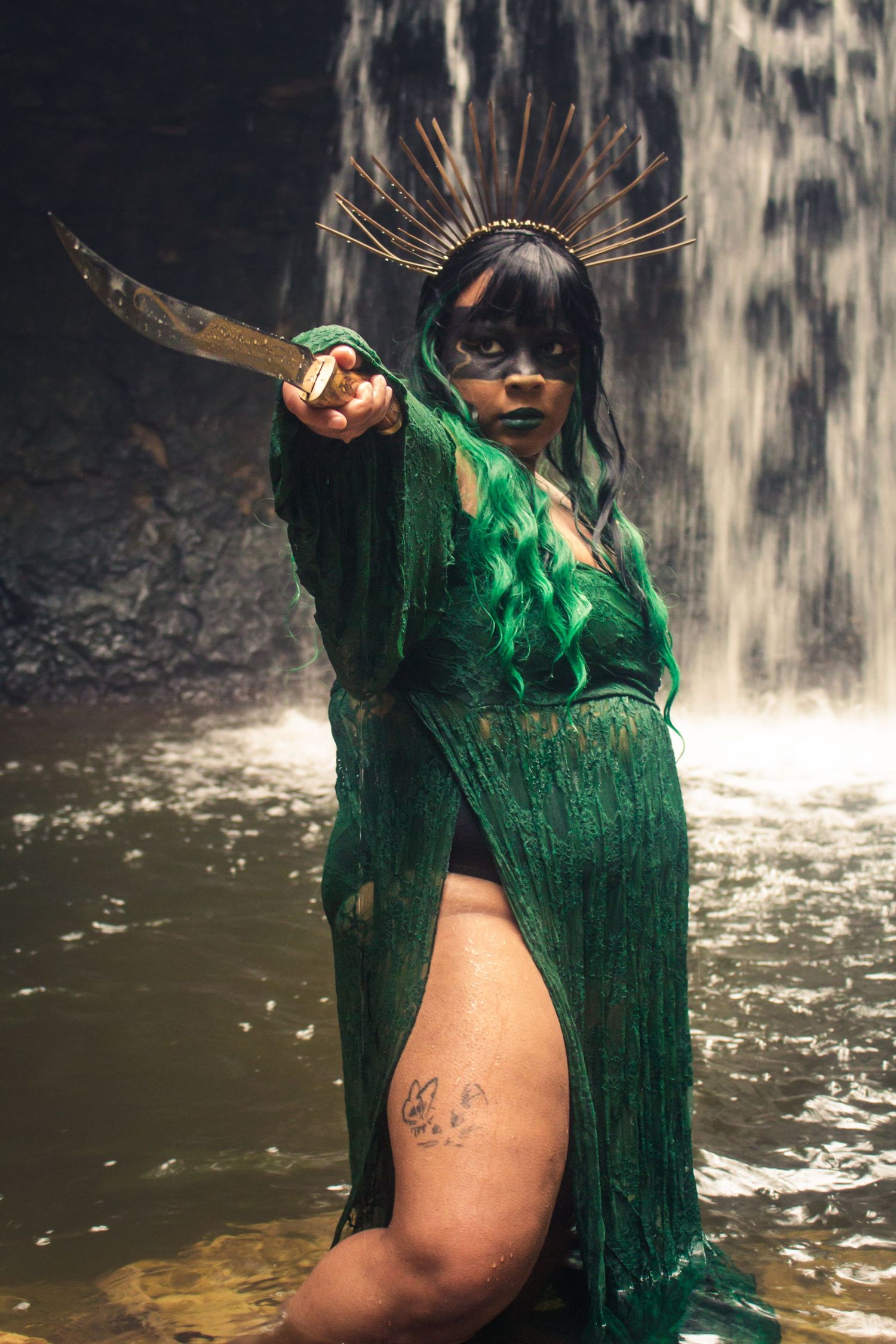 A figure in a sparkly green dress poses dramatically in front of a cascading waterfall.