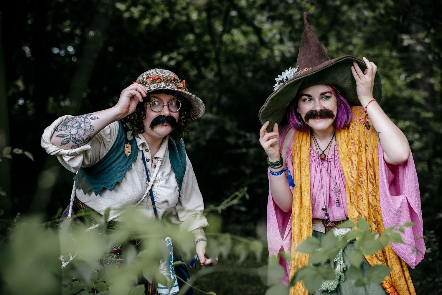 Two people in whimsical wizard costumes with fake mustaches and colorful hats pose outdoors among green foliage.