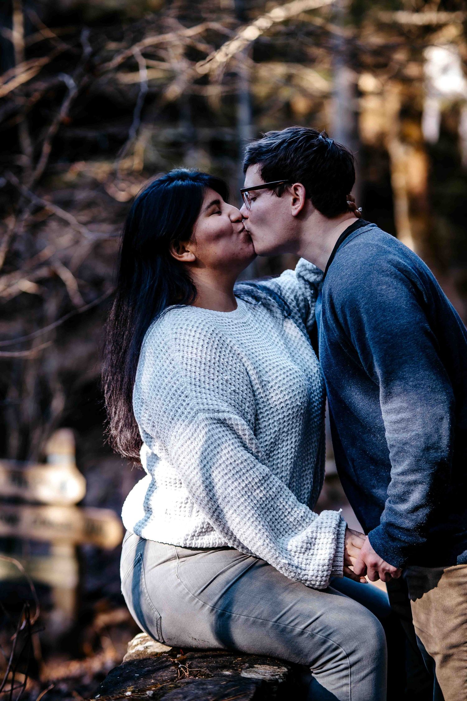 A couple shares a romantic kiss outdoors in a forest setting, wearing cozy sweaters in cool weather.
